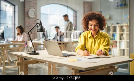 Portrait of Biracial Active Young Woman Working on Laptop in Busy Office. Biracial Female Manager smiling while Taking Notes one a Notebook From Her Laptop . People Using Computers in the Background. Stock Photo