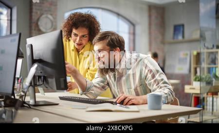Portrait of Two Creative Colleagues Collaborating and Pointing at the Computer Screen in Modern Bright Office. Biracial Young Woman Discussing Work with Caucasian Male Assistant. Stock Photo