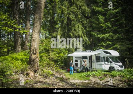 Friends standing in front of a parked camper van, looking at each other. The camper is parked by a serene lake, with trees in the background. Stock Photo