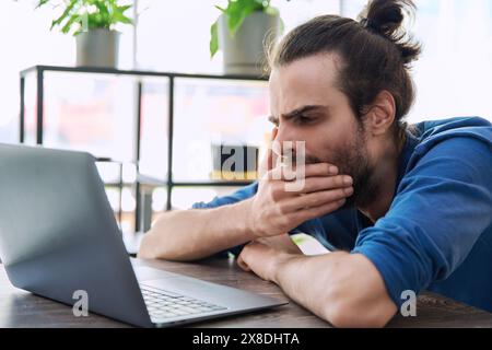 Young tired sleepy man looking at laptop, yawning covering his mouth with hand Stock Photo