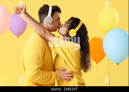 Young couple in love with headphones and balloons kissing on yellow background Stock Photo