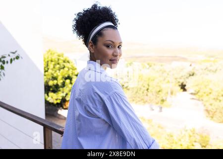 A biracial woman outdoors, wearing striped shirt on a balcony. Curly-haired with a white headband, gently smiling at camera, enjoying the view, unalte Stock Photo