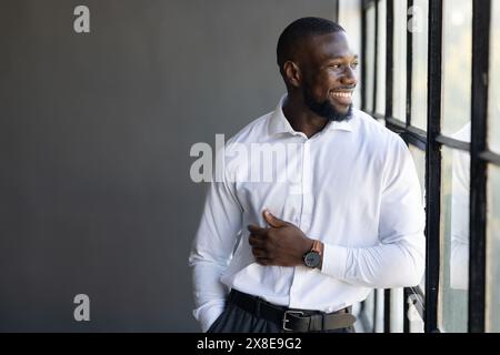 A young African American man with short hair and a beard stands by a window in an office, copy space. Wearing a white shirt, grey pants, and a black b Stock Photo