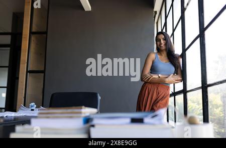 At modern office, Indian woman standing by large windows, smiling and looking relaxed, copy space. She has long black hair, light skin, and is wearing Stock Photo