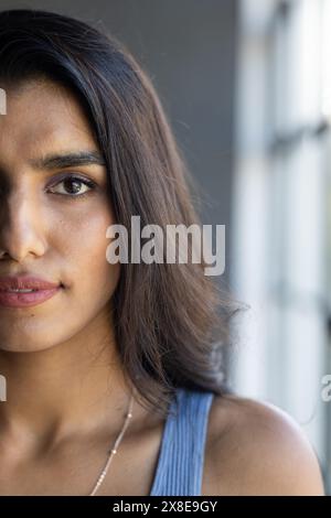At modern office, young Indian woman with long dark hair standing near window. Wearing blue top and delicate necklace, looking thoughtful and gazing o Stock Photo
