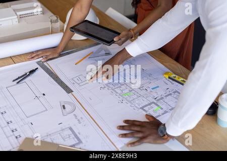 Diverse team working on architectural plans in office, focusing on blueprints. African American male pointing at blueprint while indian female team le Stock Photo