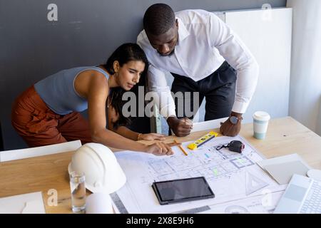 Diverse team working on architectural plans in office, focusing on blueprints. Young African American male colleague and indian female team leader dis Stock Photo