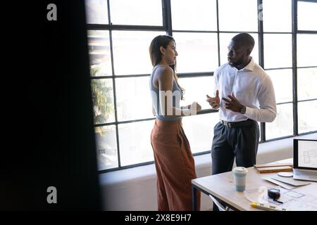 At modern office, diverse team discussing work near large windows, copy space. Young African American male with short hair and beard talking to young Stock Photo