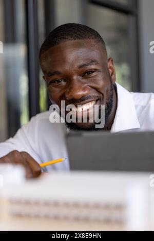 In a modern office, a young African American man smiles, holding a pencil and a tablet. Short black hair, beard, and white shirt, creating focused and Stock Photo