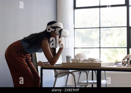 indian woman wearing virtual reality headset leaning over table in office. She is looking at architectural model with thoughtful expression, wearing s Stock Photo