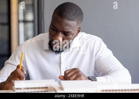 Young African American male with short hair and beard working in modern office. Wearing white shirt and watch, holding pencil and writing on paper, lo Stock Photo