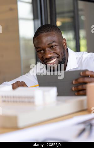 At modern office, young African American man with short black hair and beard smiles. Wearing white shirt and sitting at a desk with office supplies, h Stock Photo