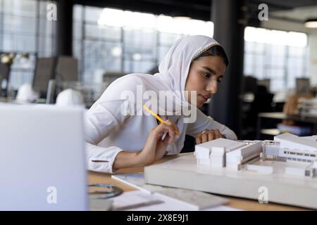 In a modern office, Indian woman in white hijab works on architectural model. She is holding pencil and focusing on details of model in office setting Stock Photo