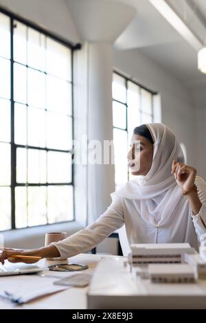 At modern office, Indian woman in white hijab and blouse works at desk, copy space. She is looking at architectural models and papers, pencil in hand, Stock Photo