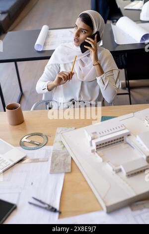 At modern office, young Indian woman wearing hijab talking on phone. She is holding a pencil and sitting at a desk with architectural drawings and a m Stock Photo