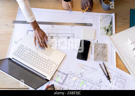 Diverse team working on architectural plans at desk with various tools. indian team leader using laptop, African American team member holding pencil n Stock Photo