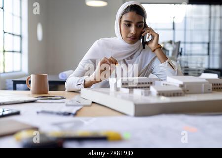 At modern office, young Indian woman wearing white hijab working on models. She is talking on phone while holding pencil, with various drawings and to Stock Photo