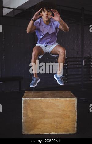 Young biracial man fitness enthusiast at gym performing box jumps. Surrounded by gym equipment and dark background, focusing on his workout with inten Stock Photo