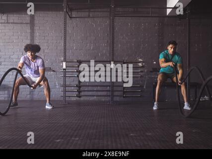 Two young biracial men are working out with battle ropes at gym with diverse men, copy space. They are in spacious, industrial-style gym with brick ba Stock Photo