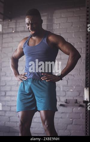 Strong and fit African American man at gym wearing blue tank top and shorts. He is standing with hands on hips against brick background, looking deter Stock Photo