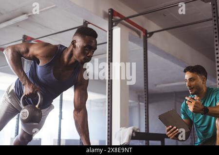 At gym, diverse men working out: African American with kettlebell, another with a tablet. Gym environment with workout equipment and supportive atmosp Stock Photo