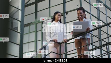 Image of notification bars over diverse female coworkers talking and using laptop on staircase Stock Photo