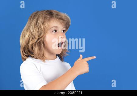 Surprised child looking away, pointing finger on isolated studio background. Shocked kid pointing to copyspace, showing promo offers, points away Stock Photo