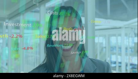 Image of texts, graphs with changing numbers, smiling biracial woman standing in office Stock Photo