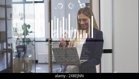 Image of graphs, loading bars over thoughtful caucasian woman standing and looking at laptop Stock Photo