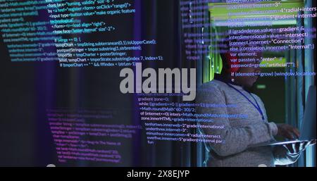 An African American man in his early thirties is working on laptop, surrounded by code on a screen Stock Photo