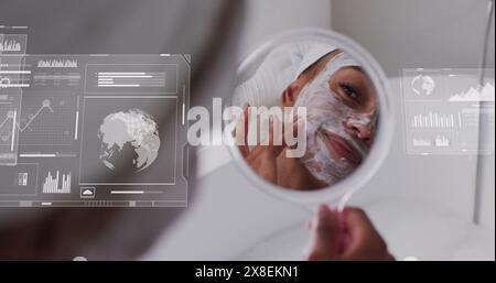 Image of graphs, globes over biracial woman looking in mirror and applying cream on face Stock Photo