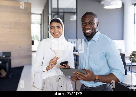 In modern office, diverse team standing, smiling at camera. African American male holding tablet, indian female team leader holding smartphone, both l Stock Photo