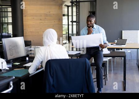 In a modern office, a young African American male holds plans with his diverse team. Talking to indian female team leader, both focused on project det Stock Photo
