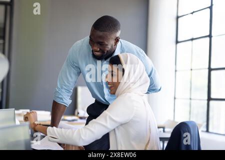 Diverse colleagues working together in office, smiling at computer screen. African American male with short black hair and indian female team leader w Stock Photo