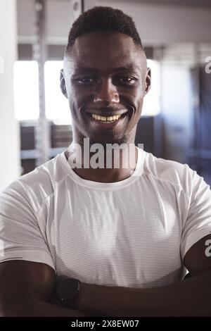 Young African American man at gym smiling confidently in white athletic shirt. He is standing in modern gym with exercise equipment and large windows Stock Photo