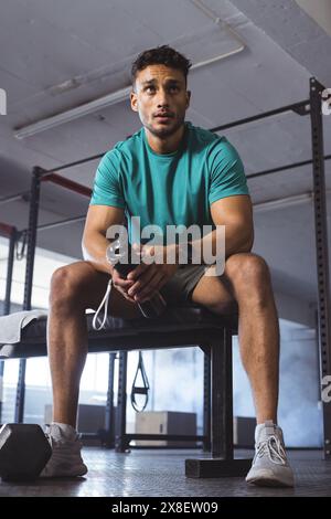At gym, biracial man sitting on gym bench holding water bottle, looking focused. Gym equipment and weights in background, creating dedicated workout a Stock Photo
