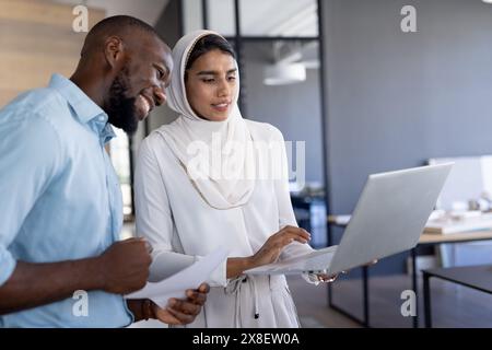 In modern office, diverse team working together, looking at laptop. African American male with short hair and beard, and indian female team leader wea Stock Photo
