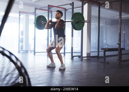 Young biracial man lifting weights at gym, looking strong and fit. He is surrounded by various gym equipment and exercising in spacious, well-lit area Stock Photo