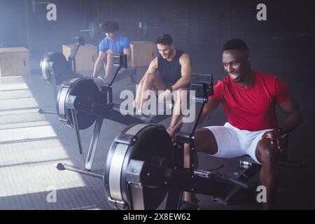 At gym, three young diverse men working out on rowing machines. They are focused on their workout in spacious, well-lit gym with various equipment in Stock Photo