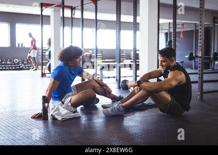 At gym, diverse men training, sitting on floor, taking break and chatting. Gym equipment and weights in background, unaltered Stock Photo