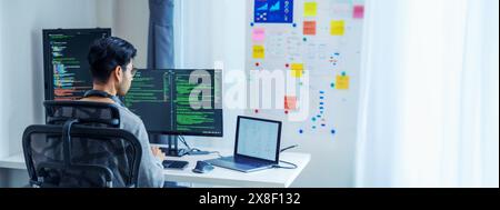 Asian software developers working on multiple screens displaying code and application diagrams in a modern office Stock Photo
