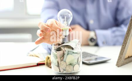 Male arm holding lightbulb over tip can full of US money Stock Photo