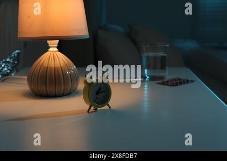 Insomnia treatment. Glass of water, pills and alarm clock on bedside table in bedroom at night, selective focus Stock Photo