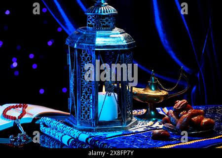 Arabic lantern, Quran, misbaha, Aladdin magic lamp, dates and folded prayer mat on mirror surface against blurred lights at night Stock Photo