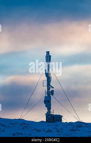 A radio tower rises tall on top of a hill covered in snow, contrasting against the white landscape. The tower looks sturdy and industrial in the winte Stock Photo