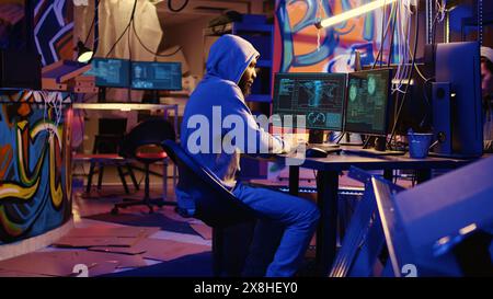 African american hacker doing computer sabotage using encryption trojan ransomware in hidden base. Hooded cybercriminal uses laptop to gain access to victims data in hideaway Stock Photo
