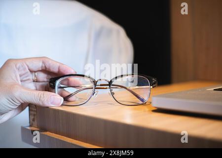 A hand holding a pair of eyeglasses near a wooden table with a laptop on it. Stock Photo