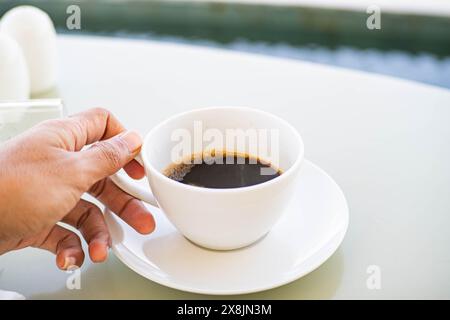 A person holding a white cup of black coffee on a white saucer, placed on a white table. The background is blurred, suggesting an outdoor setting near Stock Photo