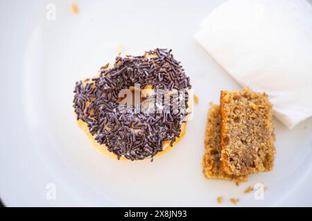 A donut with chocolate sprinkles and a piece of cake on a white plate, accompanied by a napkin. Stock Photo