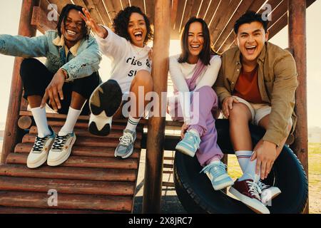 Diverse group of friends laughing and enjoying their time together outdoors on a sunny day. They are playfully posing on a wooden structure. Stock Photo